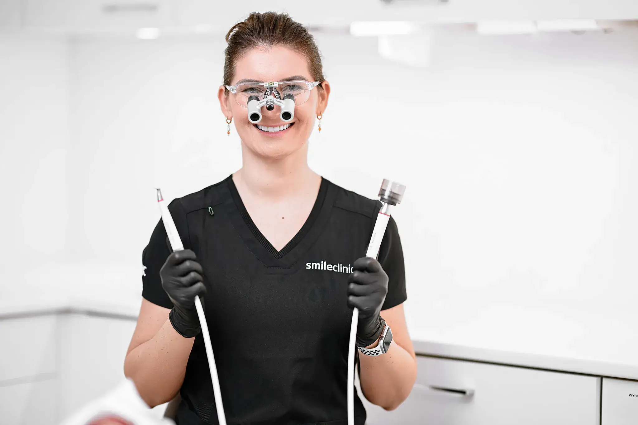 Higienistka w czarnej koszulce z logo SmileClinic trzyma narzędzia do higienizacji zębów, na białym tle, prezentując profesjonalne wyposażenie stomatologiczne.