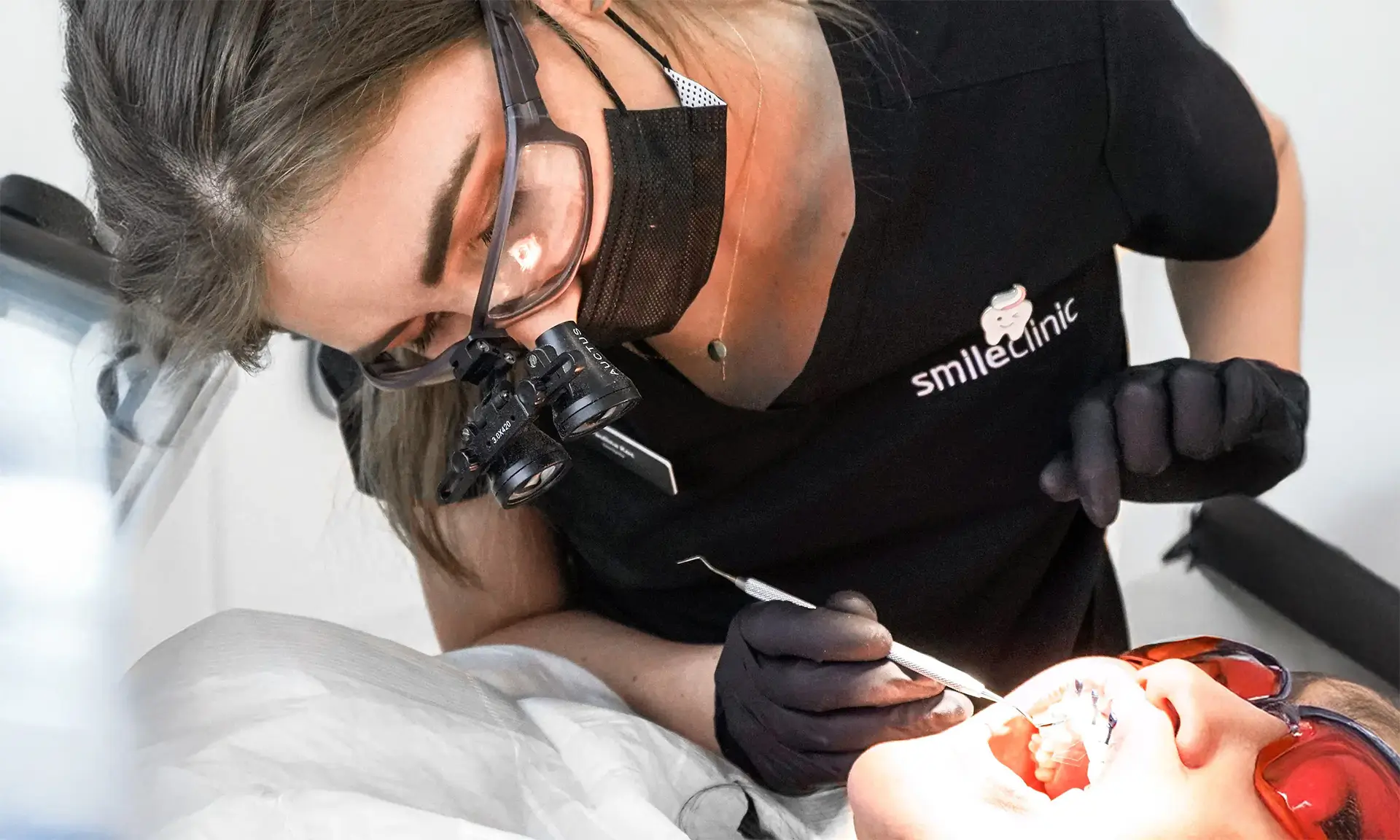 dentystka-leczenie-endodontyczne-pacjent-smileclinic-pl Dentystka w czarnej bluzie z logo SmileClinic, z maską i okularami powiększającymi, przeprowadza leczenie endodontyczne u pacjenta leżącego na fotelu, używając narzędzi stomatologicznych. Zdjęcie przedstawia profesjonalne zabiegi stomatologiczne w klinice SmileClinic, które pomagają uratować zęby przed usunięciem