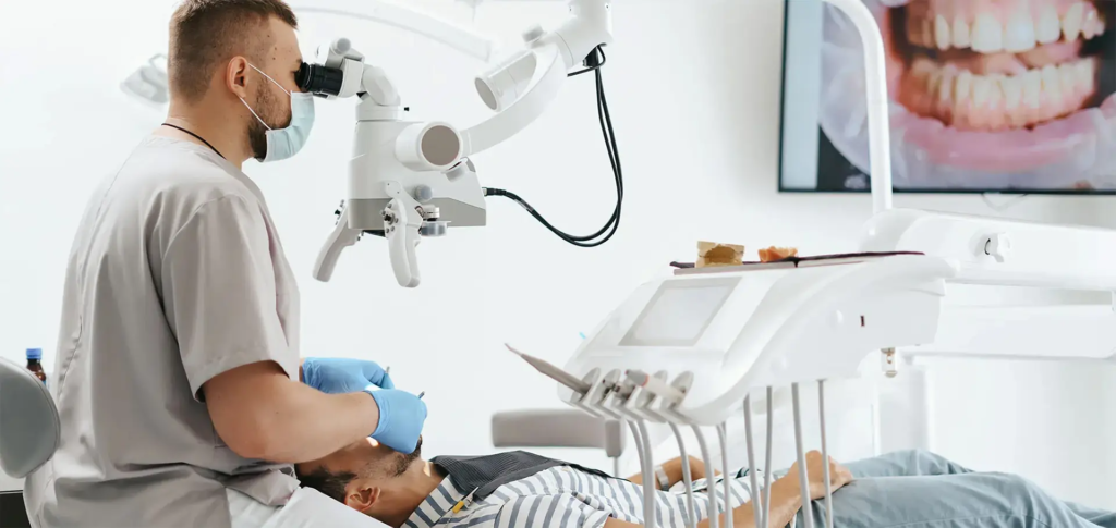 Dentystka w czarnej bluzie z logo SmileClinic, z maską medyczną i okularami powiększającymi, siedząca w gabinecie stomatologicznym na białym tle. Zdjęcie ilustruje profesjonalizm w leczeniu endodontycznym, które pozwala uratować zęby przed usunięciem, oferowanym przez klinikę SmileClinic.