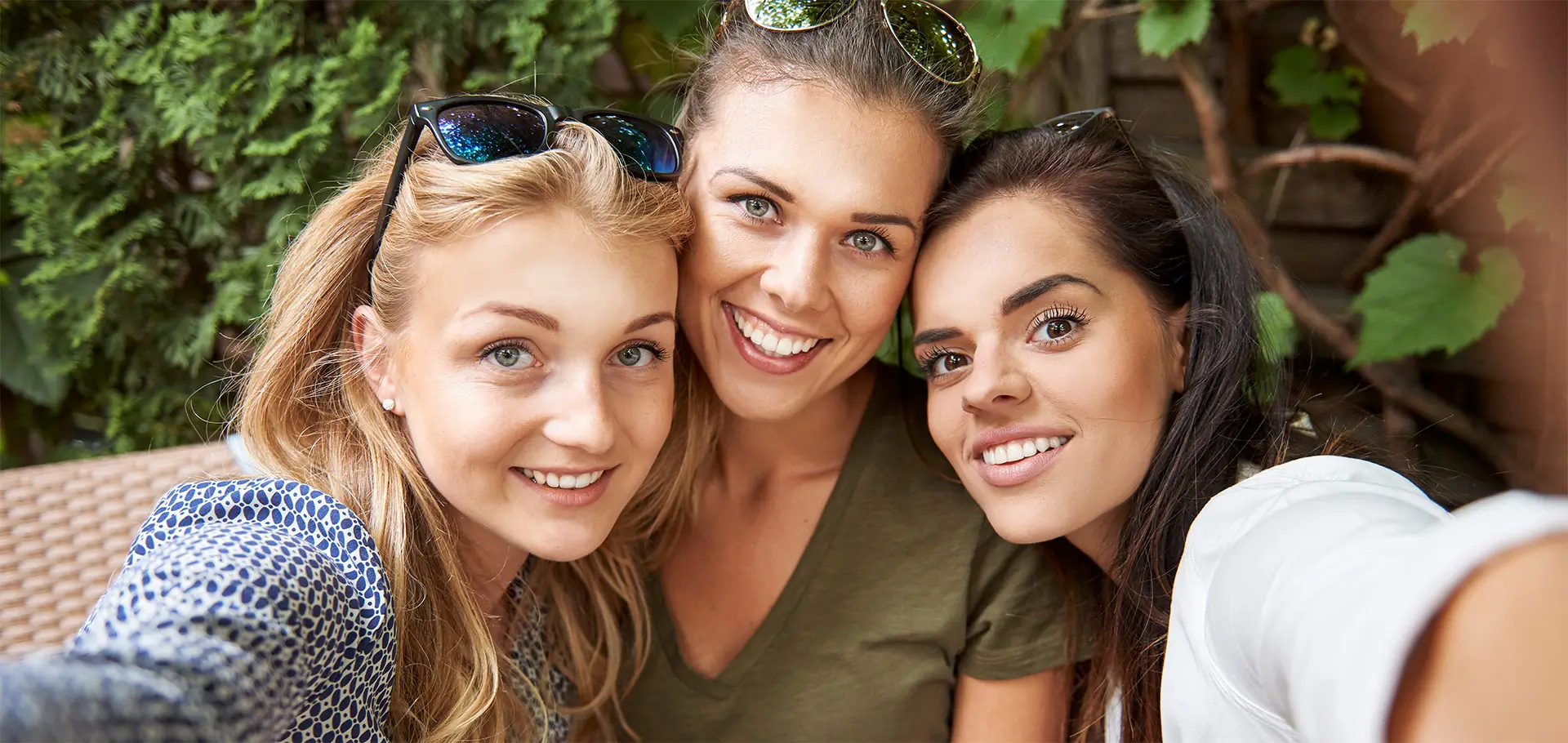 piekny-usmiech-kobiety-selfie-smileclinic-pl Trzy uśmiechnięte kobiety pozujące do selfie na zewnątrz, w otoczeniu zielonych liści. Wszystkie mają zdrowe, białe zęby i promienny uśmiech, co podkreśla temat pięknego uśmiechu i jego wpływu na zdrowie oraz samopoczucie, związany z usługami kliniki stomatologicznej SmileClinic